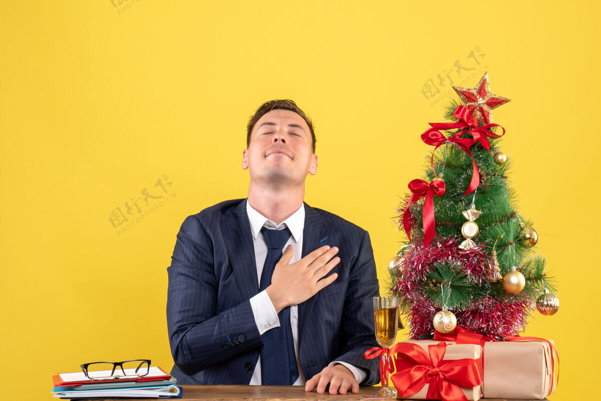 放极乐者把手放在胸前 坐在圣诞树旁的桌子旁 把礼物放在黄色的墙上前面树礼物