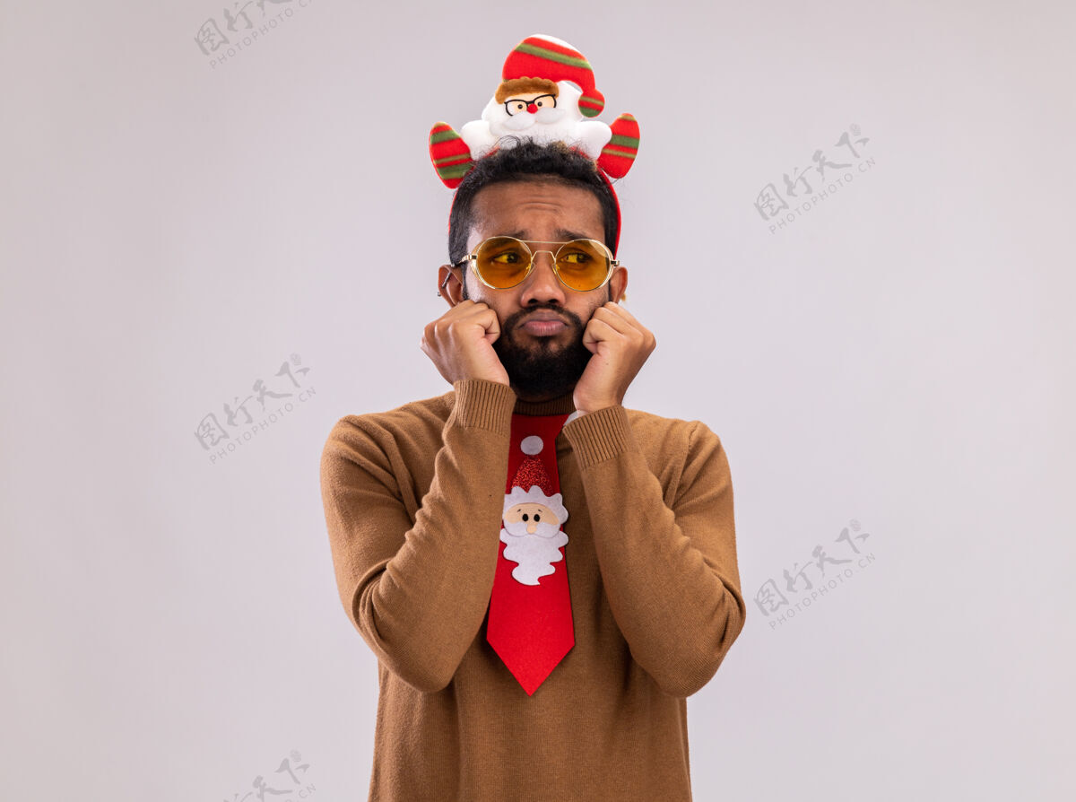 搞笑一个穿着棕色毛衣 头戴圣诞老人戒指 打着有趣的红色领带的非裔美国人站在白色背景下 一边看着一边 脸上挂着悲伤的表情头边缘表情