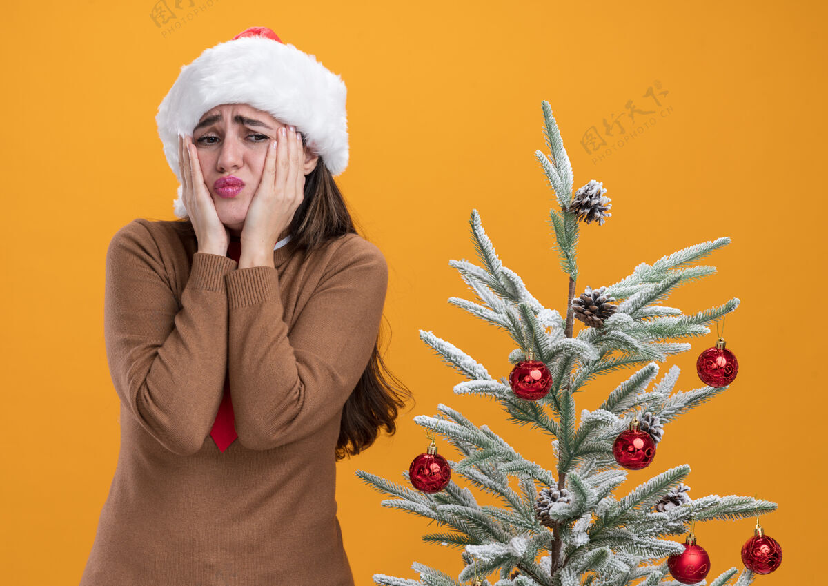 放着关心的年轻漂亮女孩戴着圣诞帽打着领带站在圣诞树旁把手放在隔离在橙色墙上的脸颊上脸颊在附近年轻
