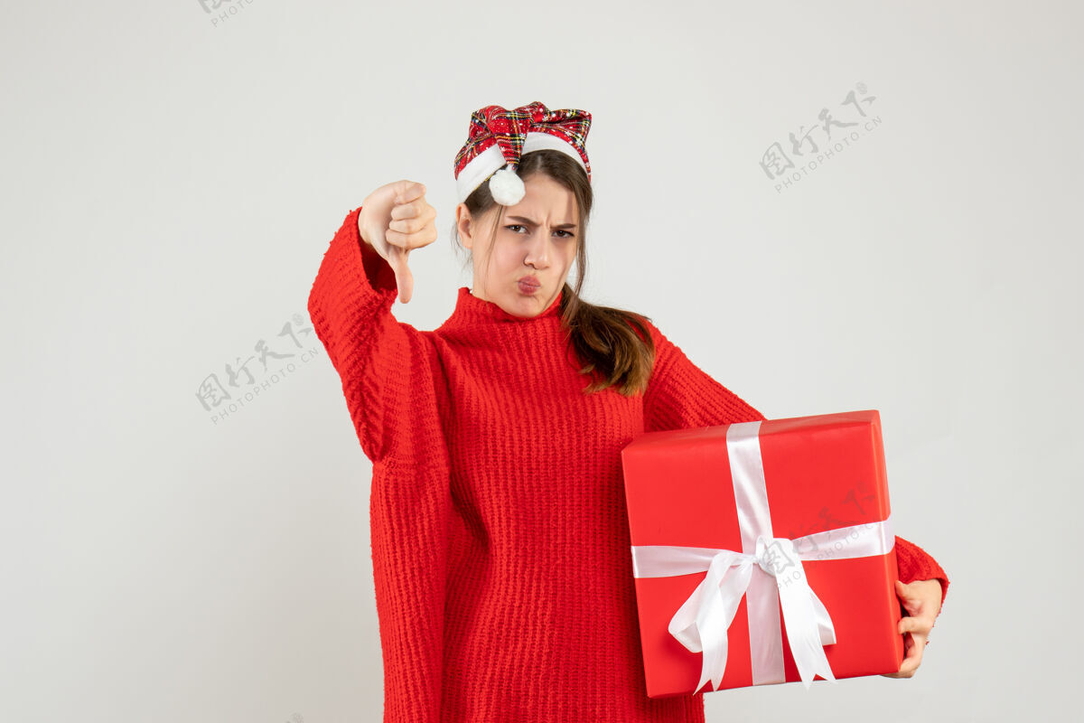 放下戴着圣诞帽拿着礼物做拇指朝下的女孩站在白色的地板上制造圣诞老人快乐