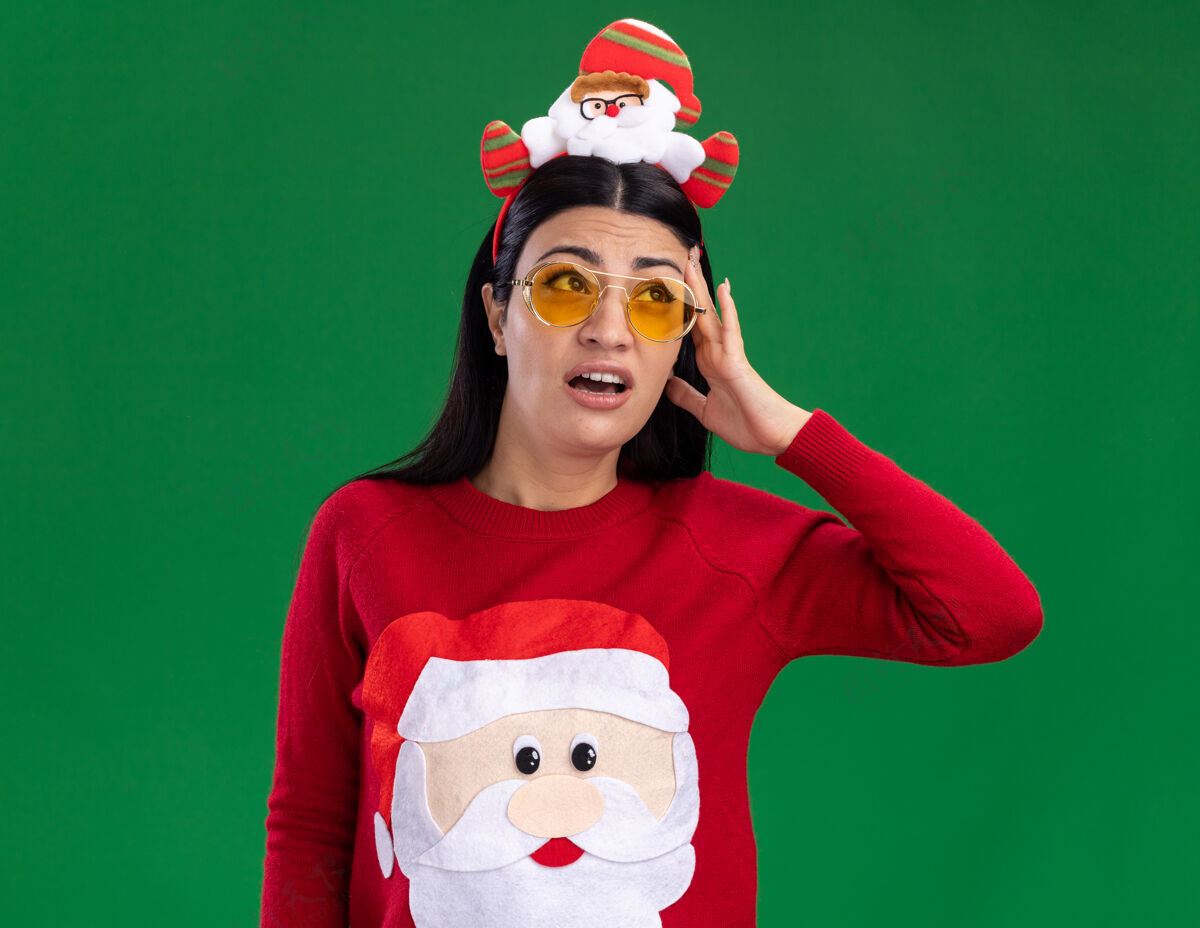 穿着困惑的年轻白人女孩戴着圣诞老人的头巾和毛衣 戴着眼镜 抬头望着绿色墙上孤立的头年轻圣诞老人毛衣