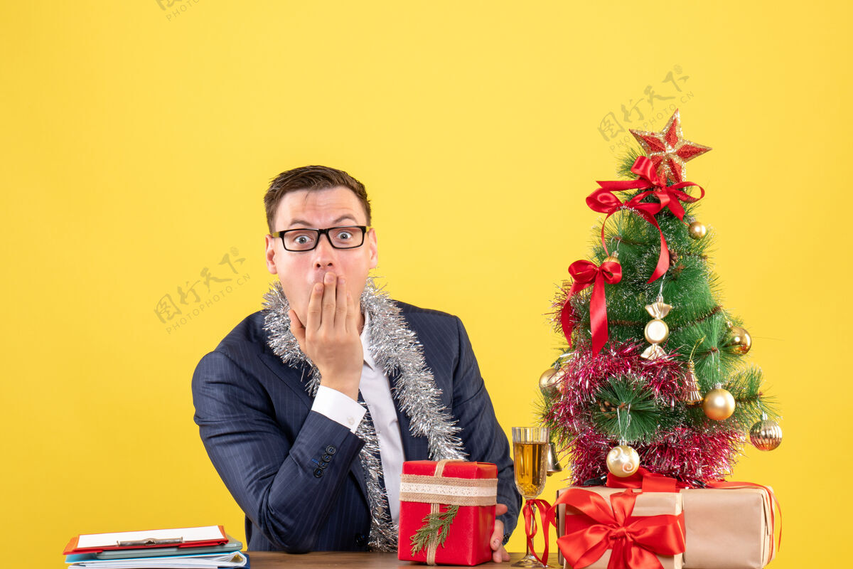 人前视图惊讶的人把手放在嘴边坐在圣诞树旁的桌子上 黄色的墙上摆着礼物肖像树礼物