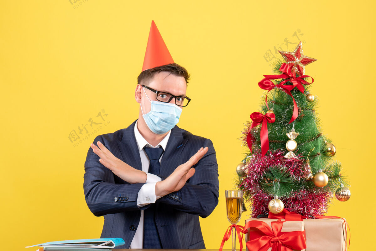 桌子商务人士交叉双手坐在圣诞树旁的桌子前 黄色墙壁上的礼物冠状病毒商人肖像