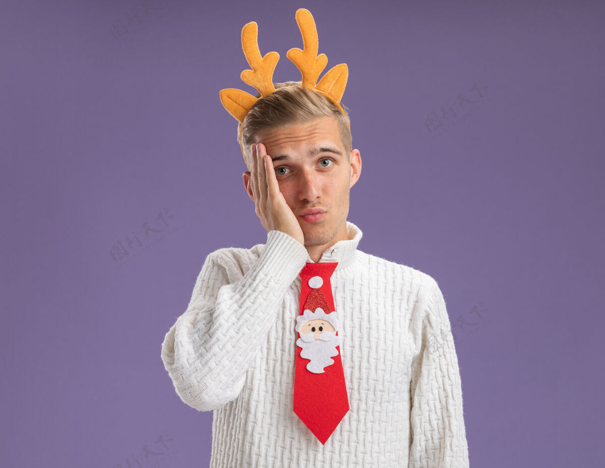 圣诞老人遗憾的是 年轻帅哥戴着驯鹿鹿角头带 打着圣诞老人的领带 看着相机 手放在脸上 在紫色的背景上 留着复制空间后悔鹿角领带