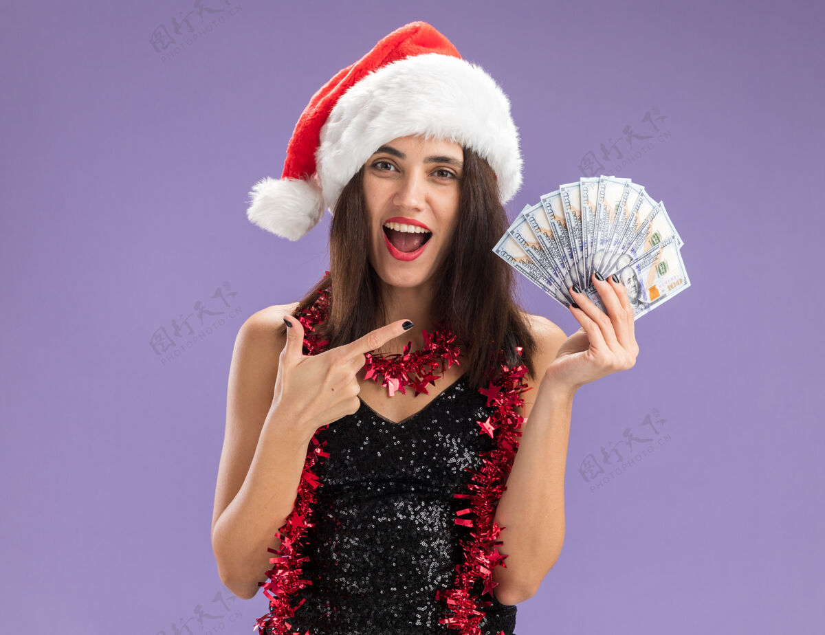 点兴奋的年轻漂亮女孩戴着圣诞帽 脖子上戴着花环 指着紫色背景上孤立的现金圣诞节女孩帽子