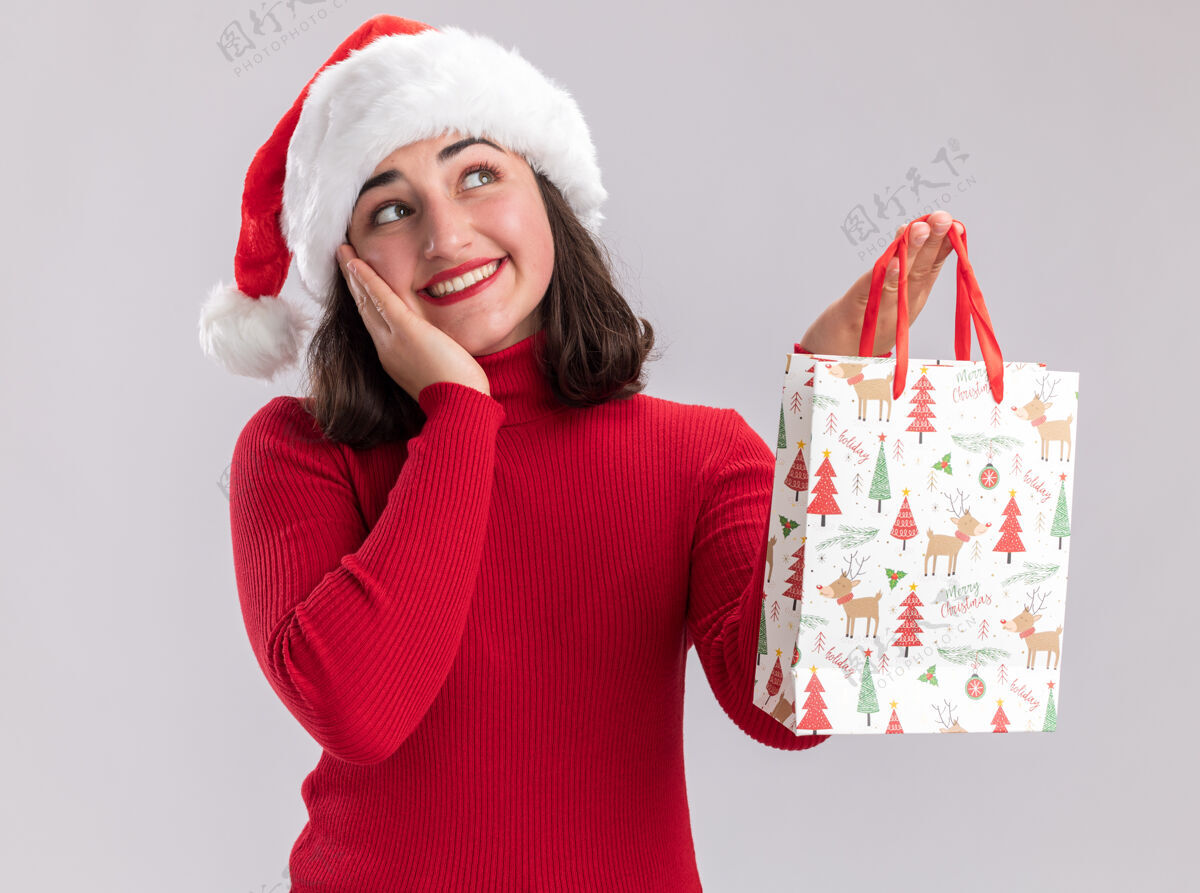 毛衣快乐的小女孩穿着红色毛衣 戴着圣诞帽 手里拿着五颜六色的圣诞礼物纸袋 站在白色的背景上 面带微笑地抬头仰望站着圣诞节包