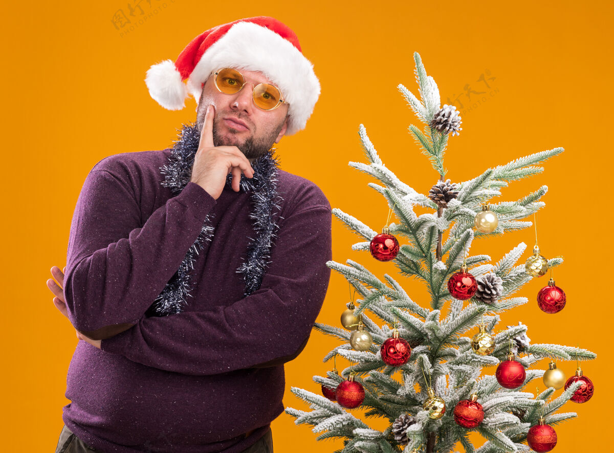 围着一位体贴的中年男子戴着圣诞帽 脖子上戴着金箔花环 戴着眼镜 站在装饰好的圣诞树旁 手放在下巴上 看着橙色背景上的相机中年体贴脖子