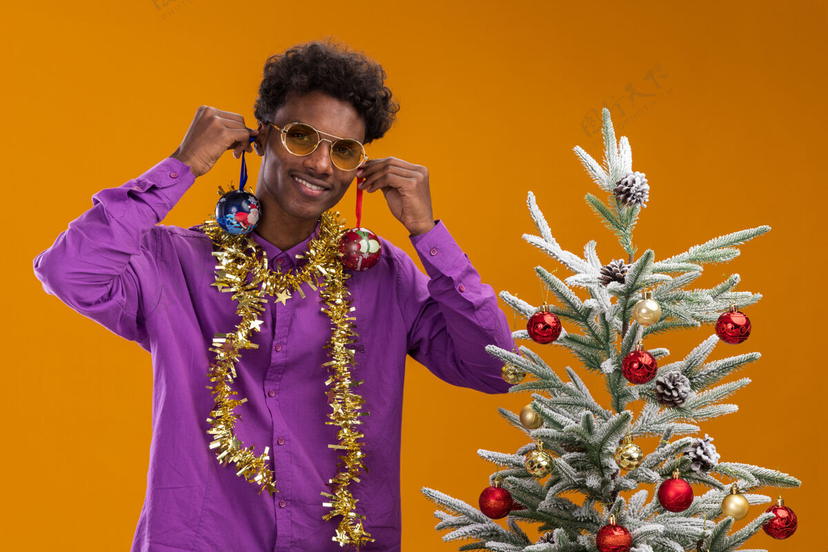 圣诞节微笑着的年轻的非裔美国人戴着眼镜 脖子上戴着金属丝花环 站在装饰过的圣诞树旁 把圣诞饰品挂在耳朵上 孤立地挂在橙色的墙上悬挂花环年轻