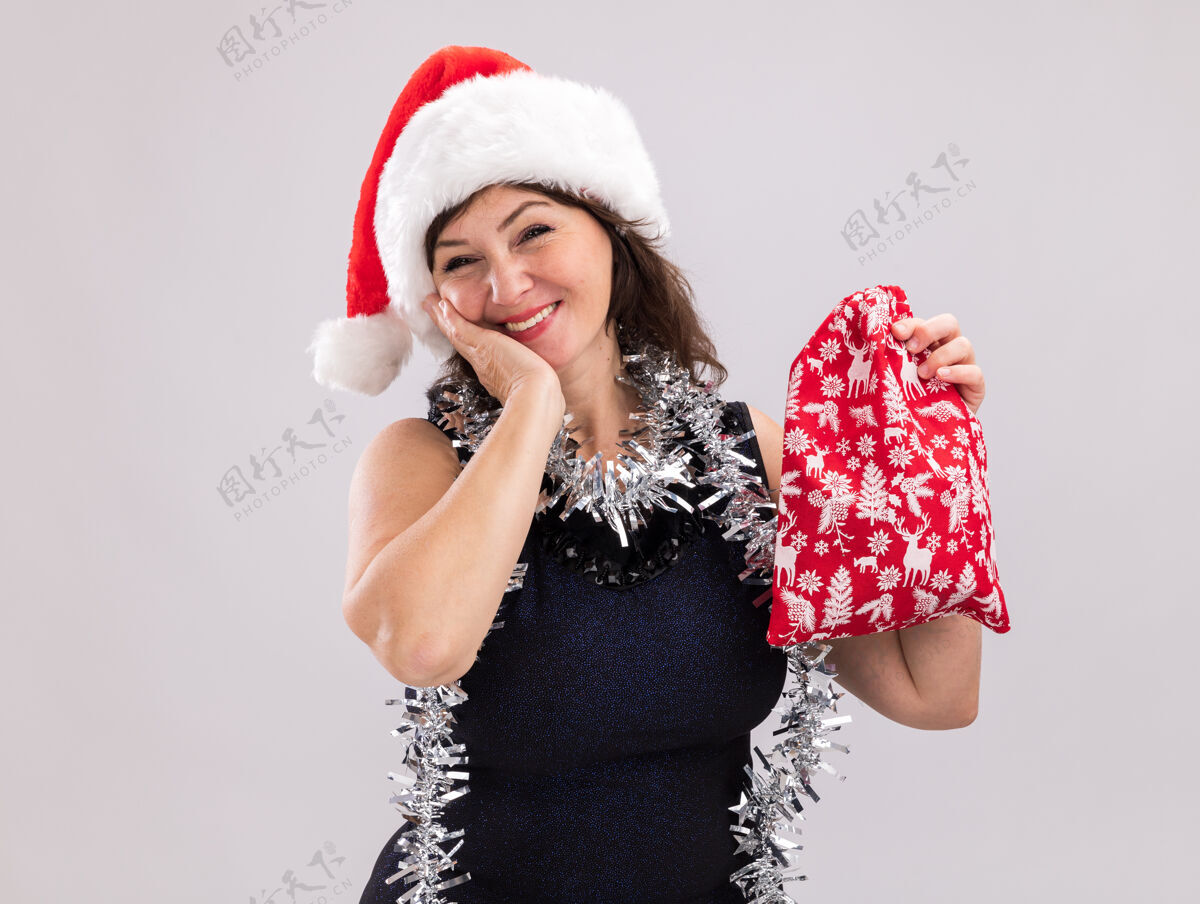 花环微笑的中年妇女戴着圣诞帽 脖子上戴着金属丝花环 手里拿着圣诞礼物袋 看着相机 手放在脸上 在白色背景上 留着复印空间金属片中年手