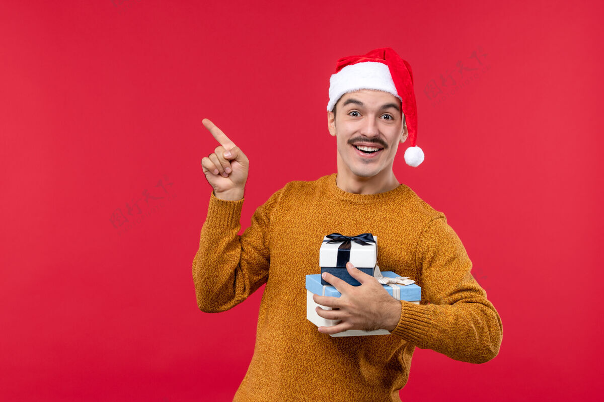 帽子红墙上拿着新年礼物的年轻人的正面图微笑年轻男性