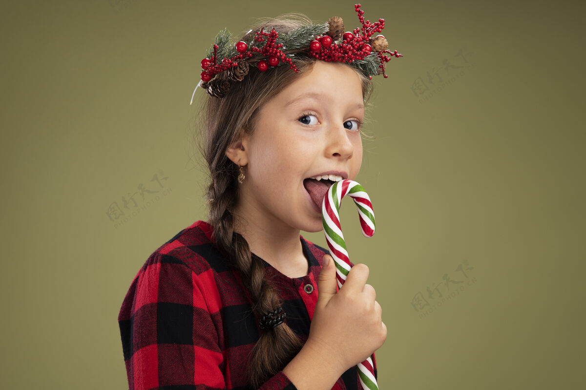 手杖小女孩戴着圣诞花圈 穿着格子裙 手持糖果手杖 站在绿色的背景下 快乐而积极地看着镜头小积极的衣服