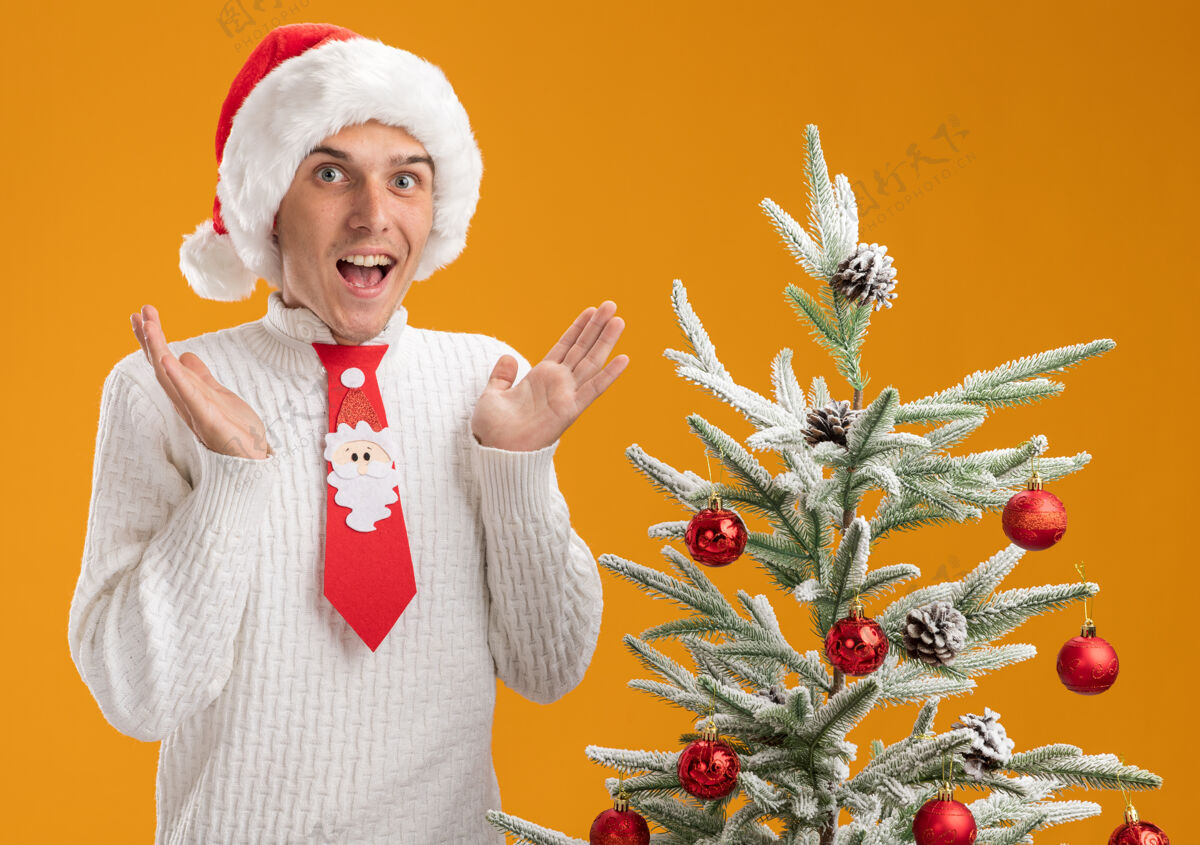 秀着令人印象深刻的年轻帅哥戴着圣诞帽 打着圣诞老人的领带 站在装饰好的圣诞树旁 两手空空的 孤零零地站在橙色的墙上戴着帽子帅气