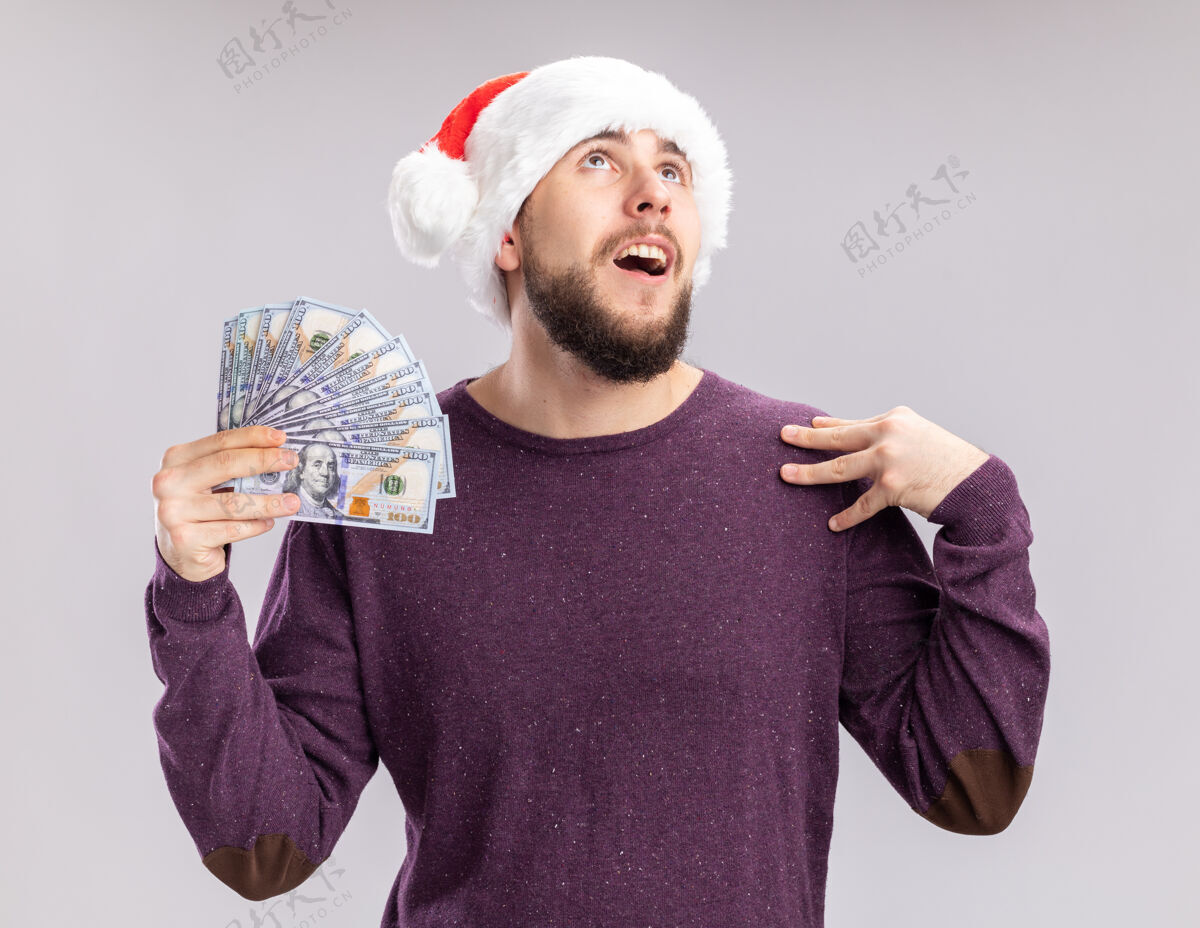 站着穿着紫色毛衣 戴着圣诞帽 手里拿着现金 站在白色背景下 高兴地微笑着抬头看的年轻人圣诞老人现金帽子