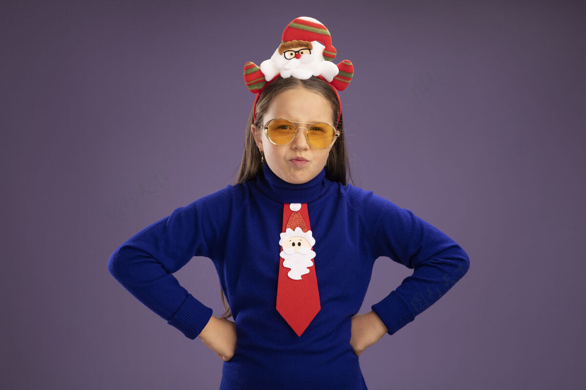 皱眉愤愤不平的小女孩穿着蓝色高领毛衣 打着红色领带 头上戴着滑稽的圣诞戒指 皱着眉头 胳膊放在臀部 站在紫色背景上 看着摄像机边缘臀部头