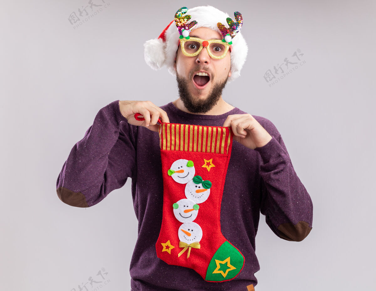 圣诞节一个穿着紫色毛衣 戴着圣诞帽 戴着滑稽眼镜 手里拿着圣诞长袜 微笑着站在白色背景下看镜头的快乐而惊讶的年轻人圣诞老人站着微笑