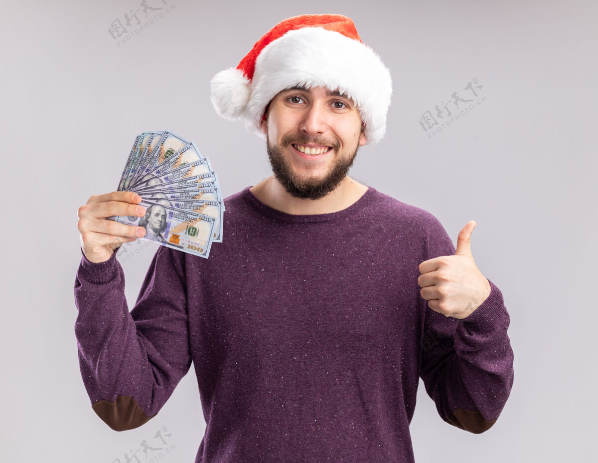 抱着一个穿着紫色毛衣 戴着圣诞帽 手里拿着现金 微笑着站在白色背景上竖起大拇指的快乐年轻人微笑年轻圣诞老人