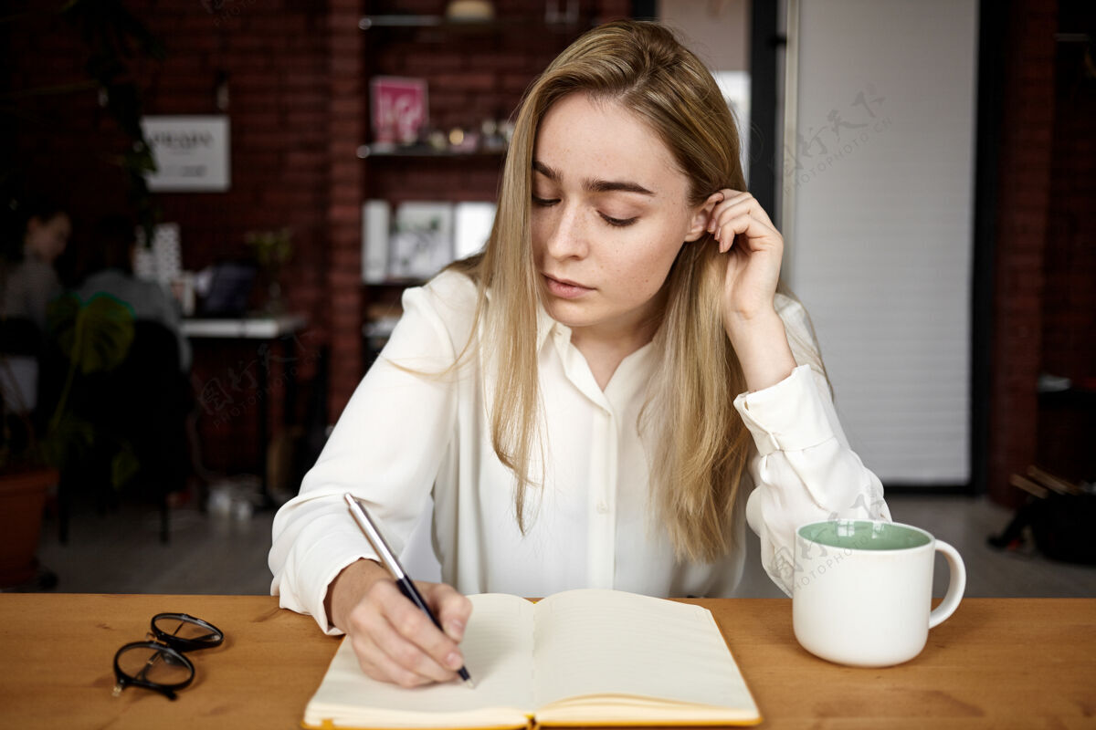 信穿着白衬衫的漂亮金发女学生在家里的工作场所做作业 在翻开的抄写本上写下 喝着茶 表情严肃专注记者日记作家