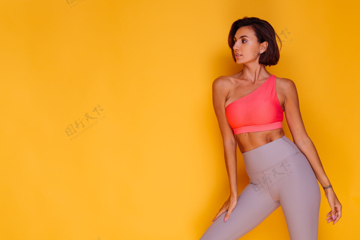 动机年轻健壮的女人穿着运动服 时髦的上衣和紧身裤 对着黄色的墙壁摆姿势锻炼健康饮食