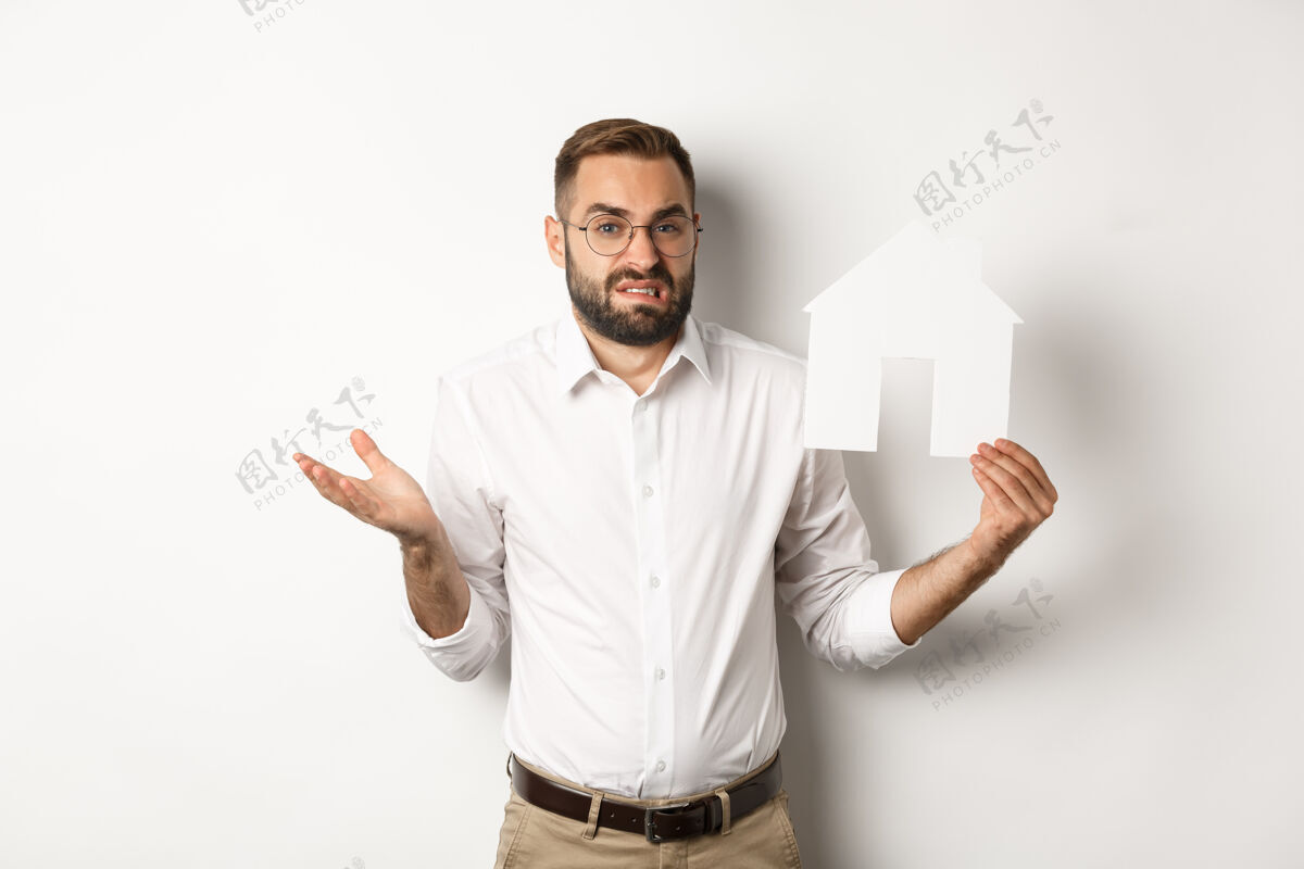 商人房地产迷茫的男子耸耸肩 展示房子的纸模型 看起来优柔寡断 站在复制空间困惑成年人时髦