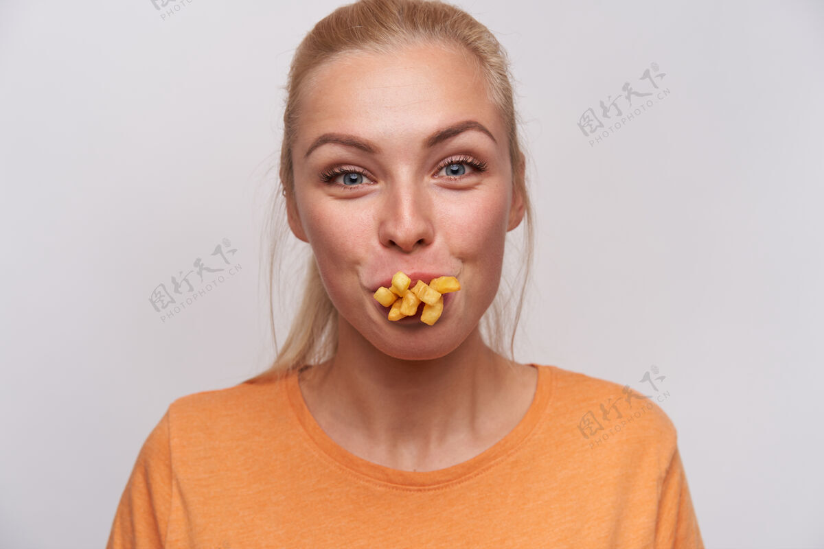垃圾有趣迷人的金发女郎的特写镜头 随意的发型 愉快地看着镜头 嘴里塞满了炸薯条 在白色背景下摆姿势 玩弄食物穿着食物享受