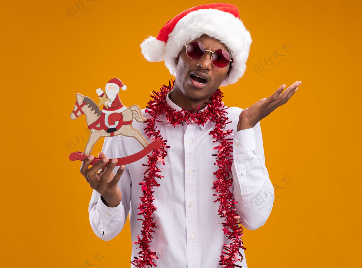 周围恼怒的美国黑人年轻人戴着圣诞帽 戴着眼镜 脖子上戴着金箔花环 把圣诞老人抱在摇摇晃晃的马俑上 看着摄像机 橙色背景上孤立着一只空手空圣诞老人恼火