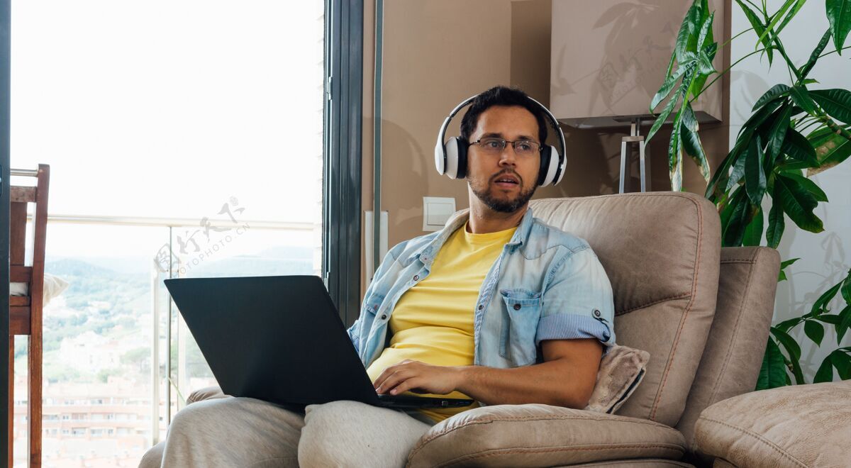 笔记本电脑一个男人用耳机和笔记本电脑跪着听音乐的水平镜头窗户男性沙发
