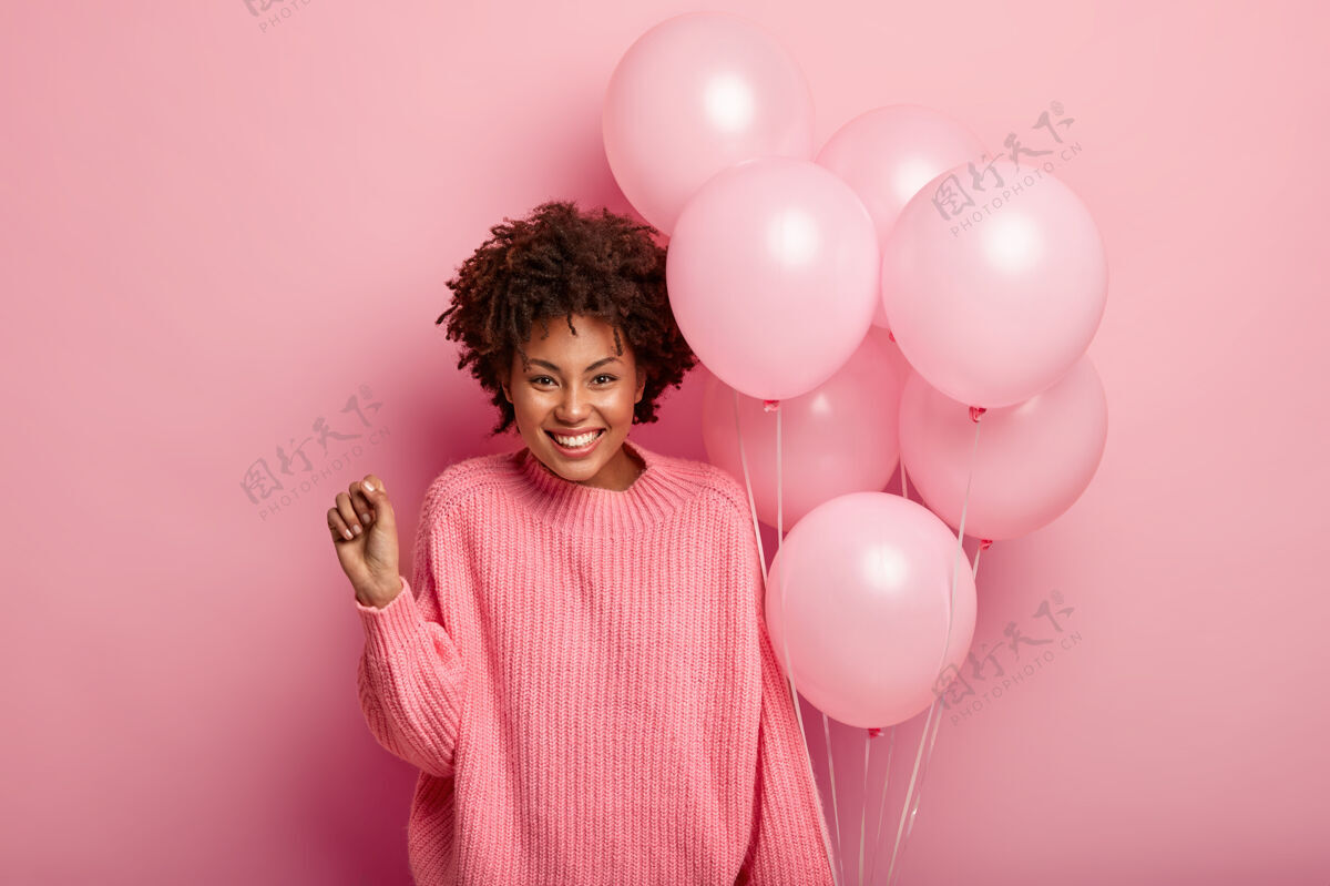 青春高兴的卷发女模特握紧拳头 穿着超大套头衫 手持气球 高兴地出席生日庆典 穿着粉色毛衣与墙壁同色牙齿快乐喜出望外