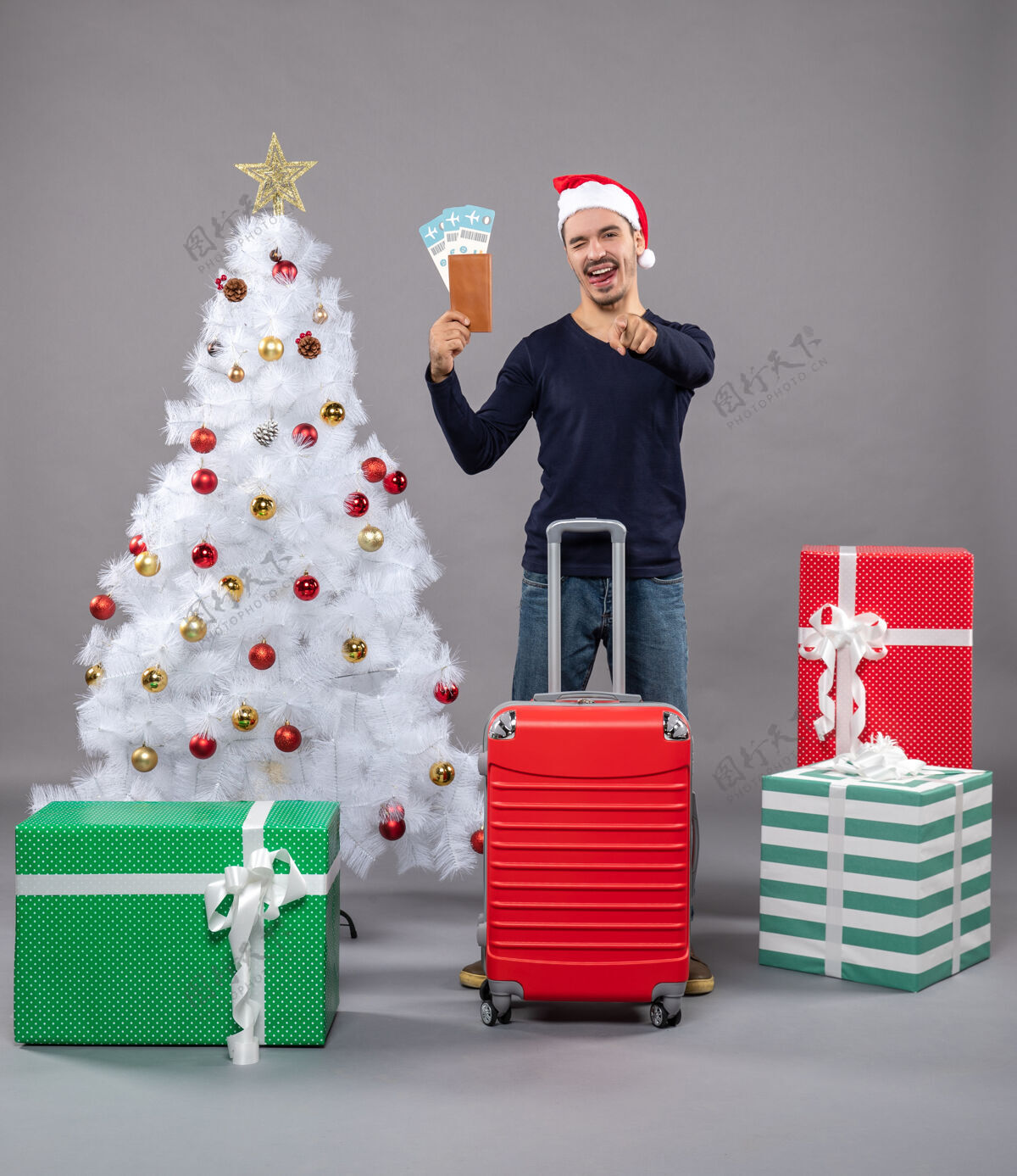 圣诞带着红色手提箱的舌头男在白色圣诞树周围展示他的旅行票和灰色的礼物展示舌头的男人红色包