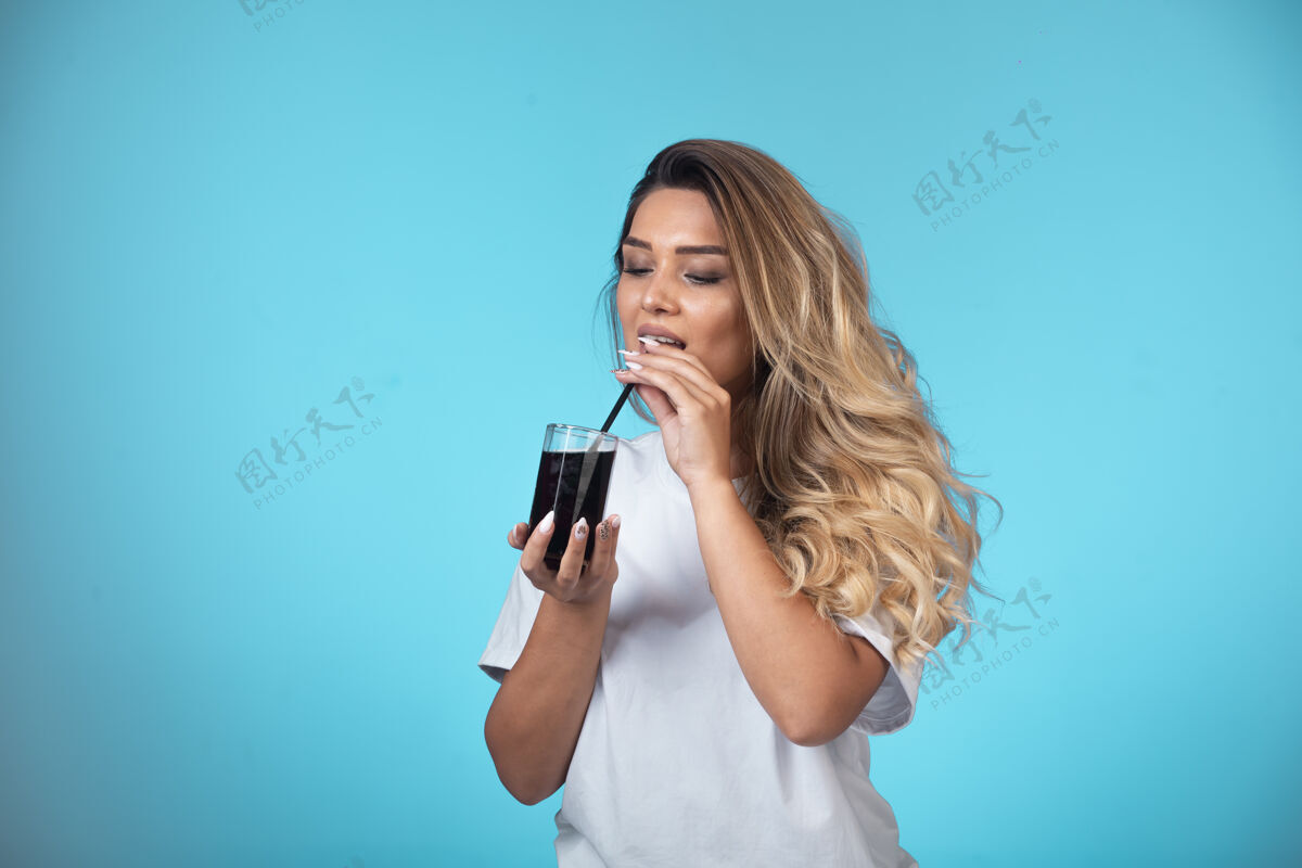 成人穿着白衬衫的年轻女孩拿着一杯黑鸡尾酒 在烟斗上检查味道摆姿势人类果汁
