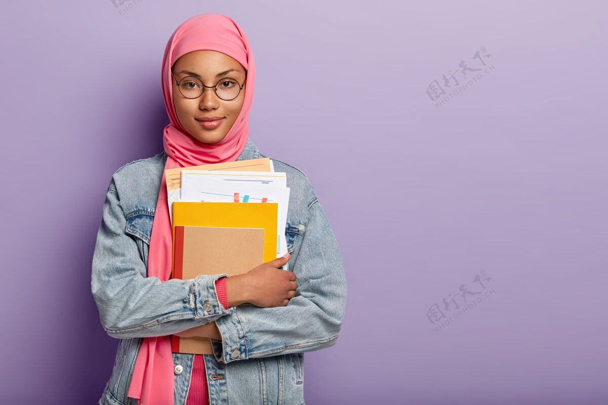 教育迷人自信的穆斯林大学生半身镜头手持笔记本 纸质文件 备课作业 戴粉色头巾 圆眼镜 牛仔服学习概念眼镜阿拉伯语夹克