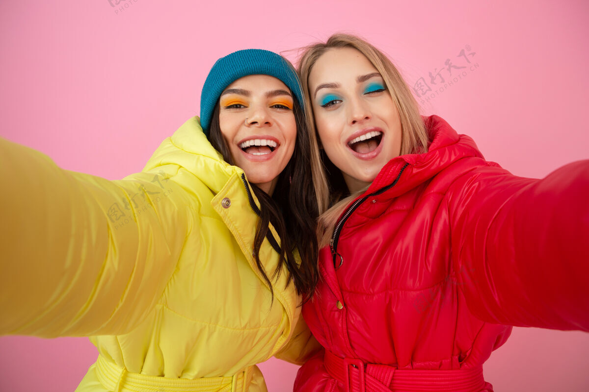 羽绒服两个迷人的女人穿着鲜艳的红黄相间的冬季羽绒服 在粉色的背景下摆着姿势 朋友们在一起玩得很开心 温暖的服装时尚潮流 自拍外套女孩衣服
