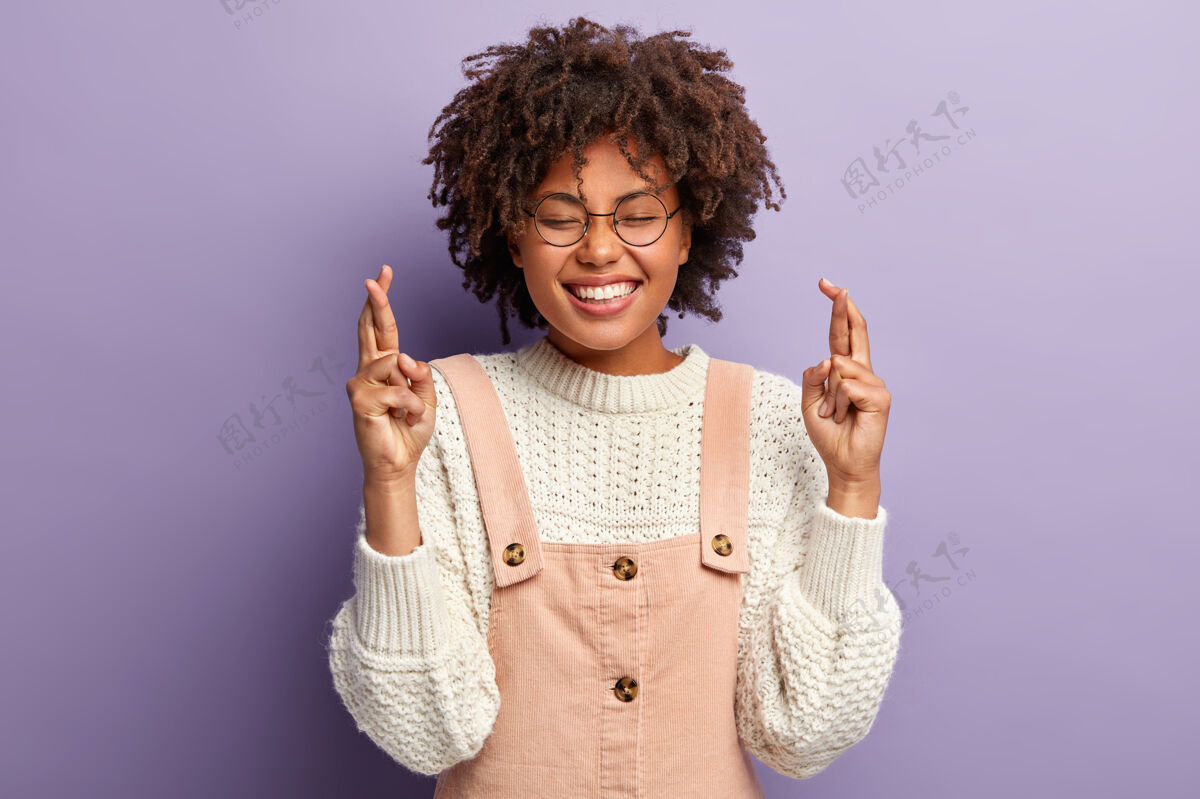 眼镜年轻开朗的女人祈祷 希望最好 笑容灿烂 穿着白色的套头衫和工作服 许下愿望 为亲人祈祷 想达到目标 孤零零地站在紫色的墙上非洲情绪希望