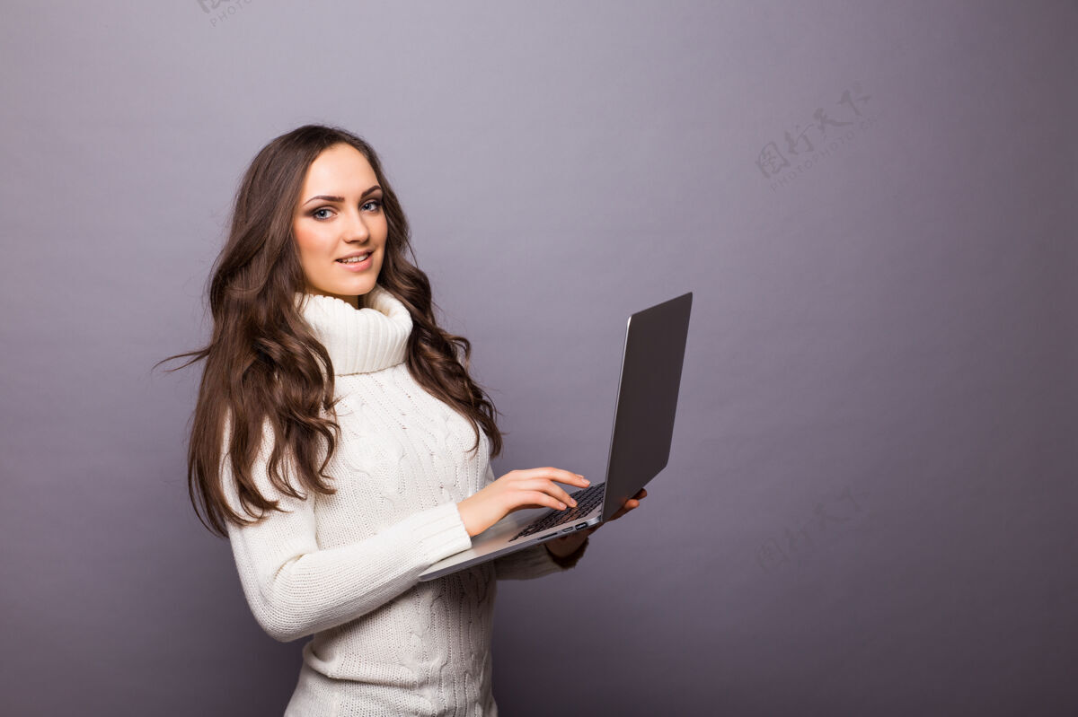 打字一个快乐的惊讶的女人站在灰色的墙上 笔记本电脑被隔离休闲聊天连接