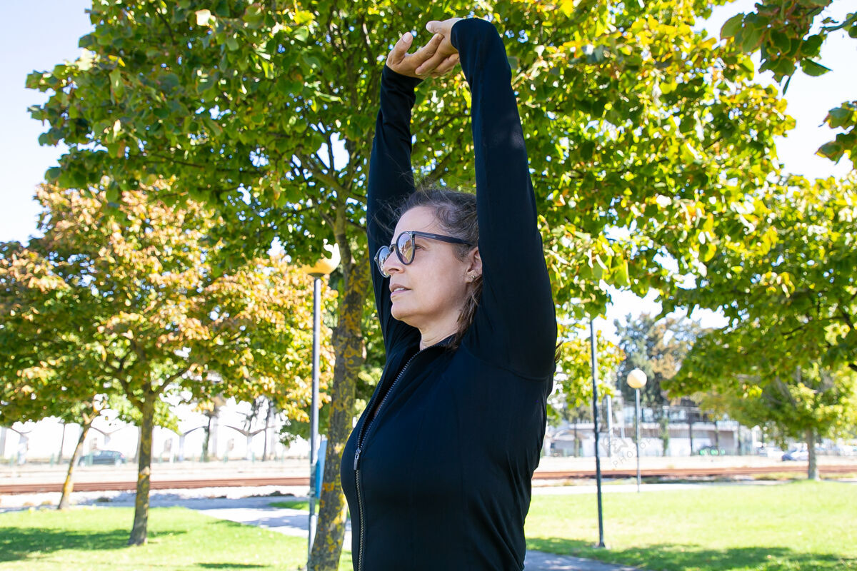 慢跑专注于运动的中年女性伸展身体 举起双手 在公园里运动时看向别处健康或积极的生活方式概念高加索中运动