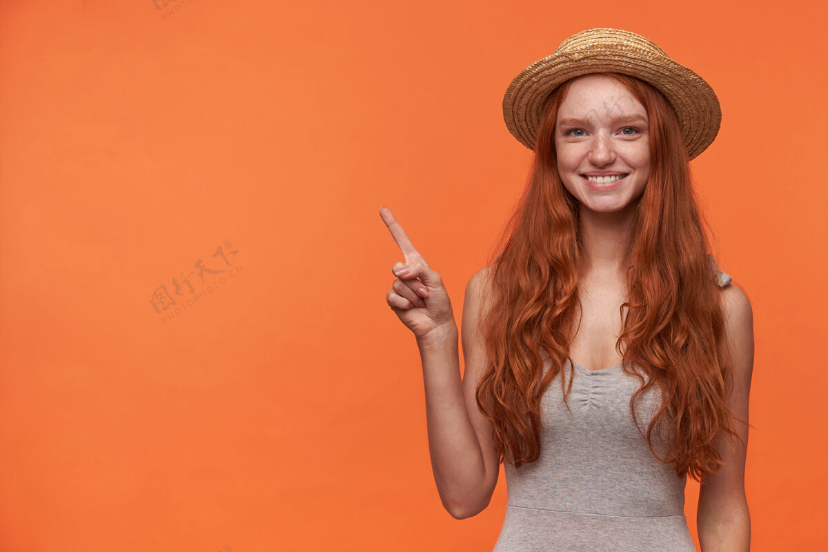 长摄影棚拍摄了一个快乐的年轻红发女子 长长的卷发指向一边 食指抬起 看着相机 开心地微笑着 穿着休闲服隔离在橙色背景下微笑食指指向