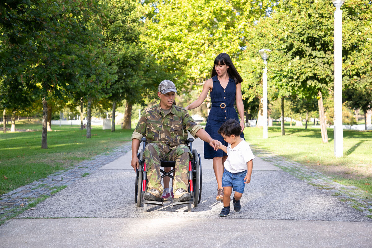 白种人严肃的军人父亲和家人坐在轮椅上散步穿着迷彩服的白人中年父亲牵着儿子的手和漂亮的妻子交谈退伍军人和残疾概念走路户外帽子