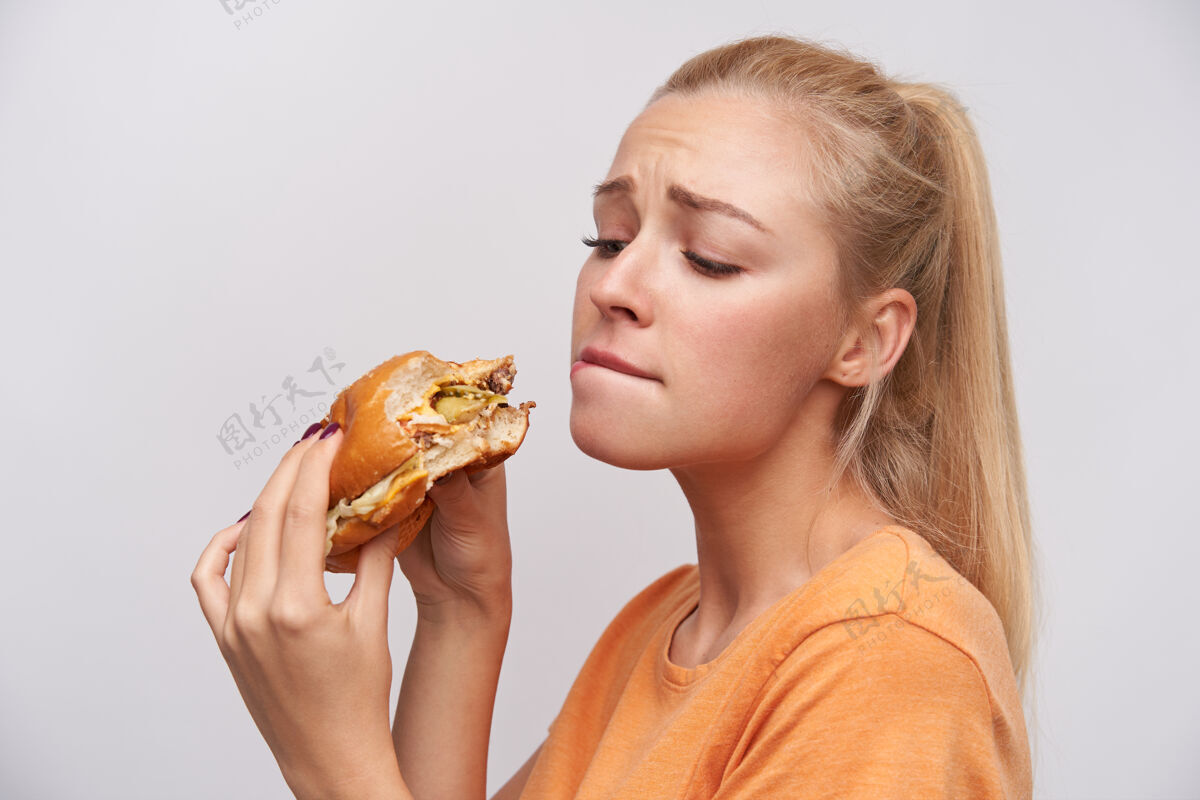 漂亮一幅年轻可爱的金发碧眼长发女性的肖像 马尾辫的发型让汉堡包高举双手 面不改色地盯着汉堡包 咬着屁股 在白色背景下皱眉天然汉堡女性