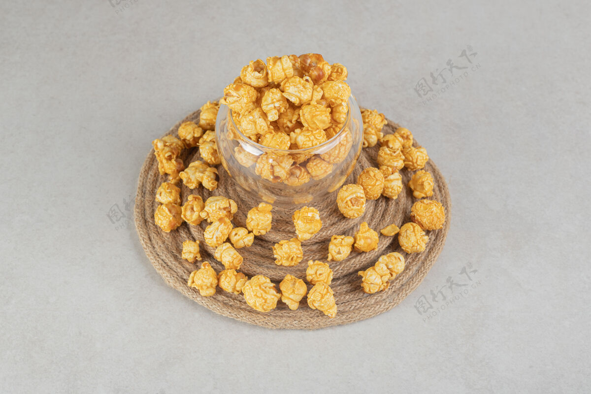 香料一个小玻璃碗在一个编织的三角架上 周围是涂有焦糖的爆米花的大理石垃圾食品美味爆米花