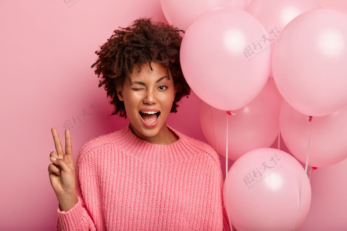 乐观深色皮肤的女模特眨着眼睛 表现出和平的姿态 穿着粉色的套头衫 带着气球 在好友圈里玩得很开心 与玫瑰色的墙壁隔绝青春 庆祝的概念满意高兴青春