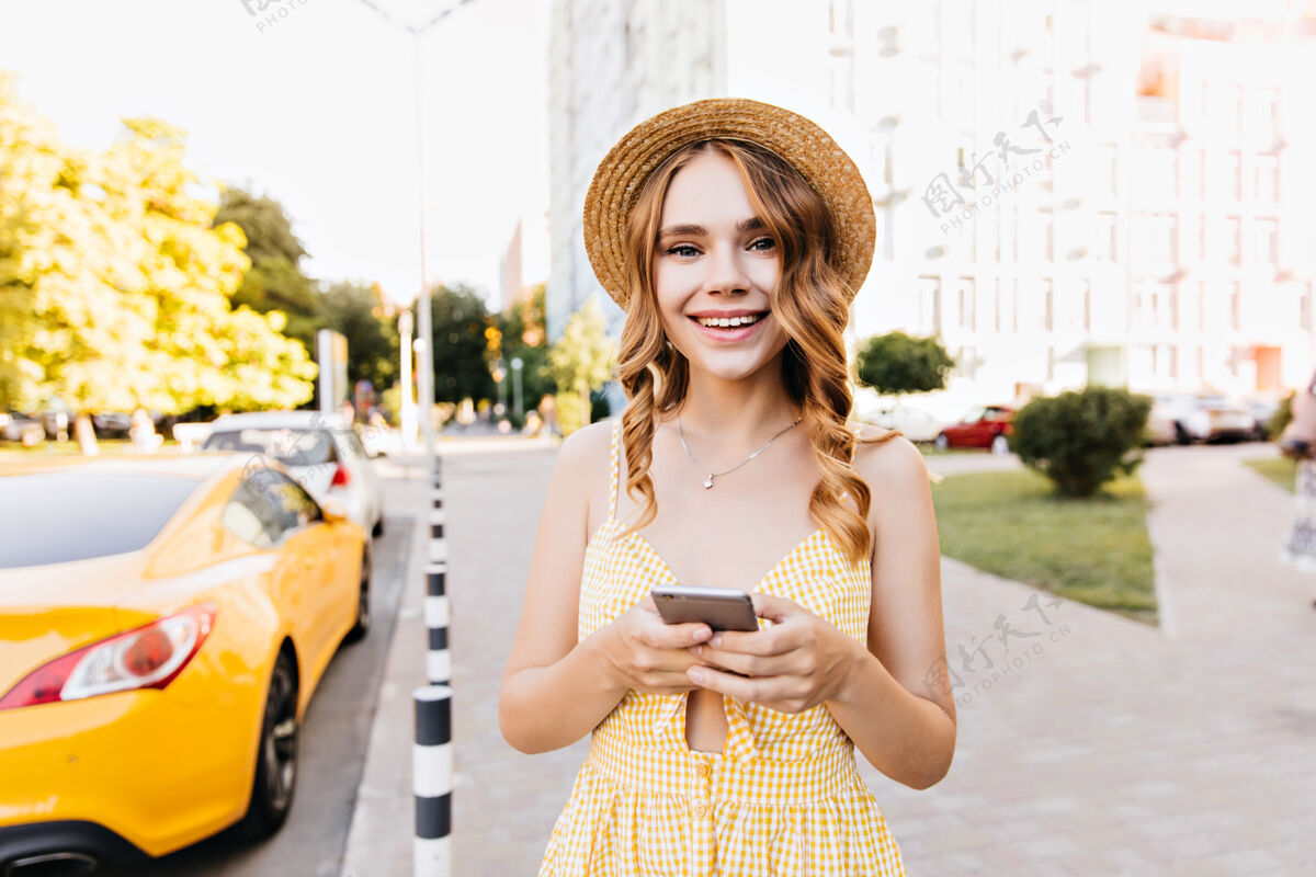 乐趣穿着复古黄色连衣裙的梦幻女孩 在散步时表达积极的情绪一个漂亮的女人 卷发 手持智能手机灵感周末草帽