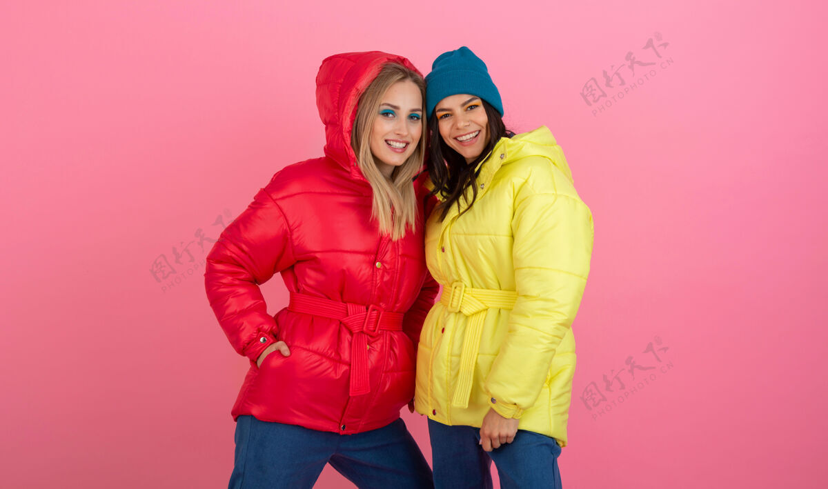 温暖的衣服两个迷人的女孩在粉红色的背景下 穿着鲜艳的红色和黄色的彩色冬季羽绒服摆姿势温暖粉色女人