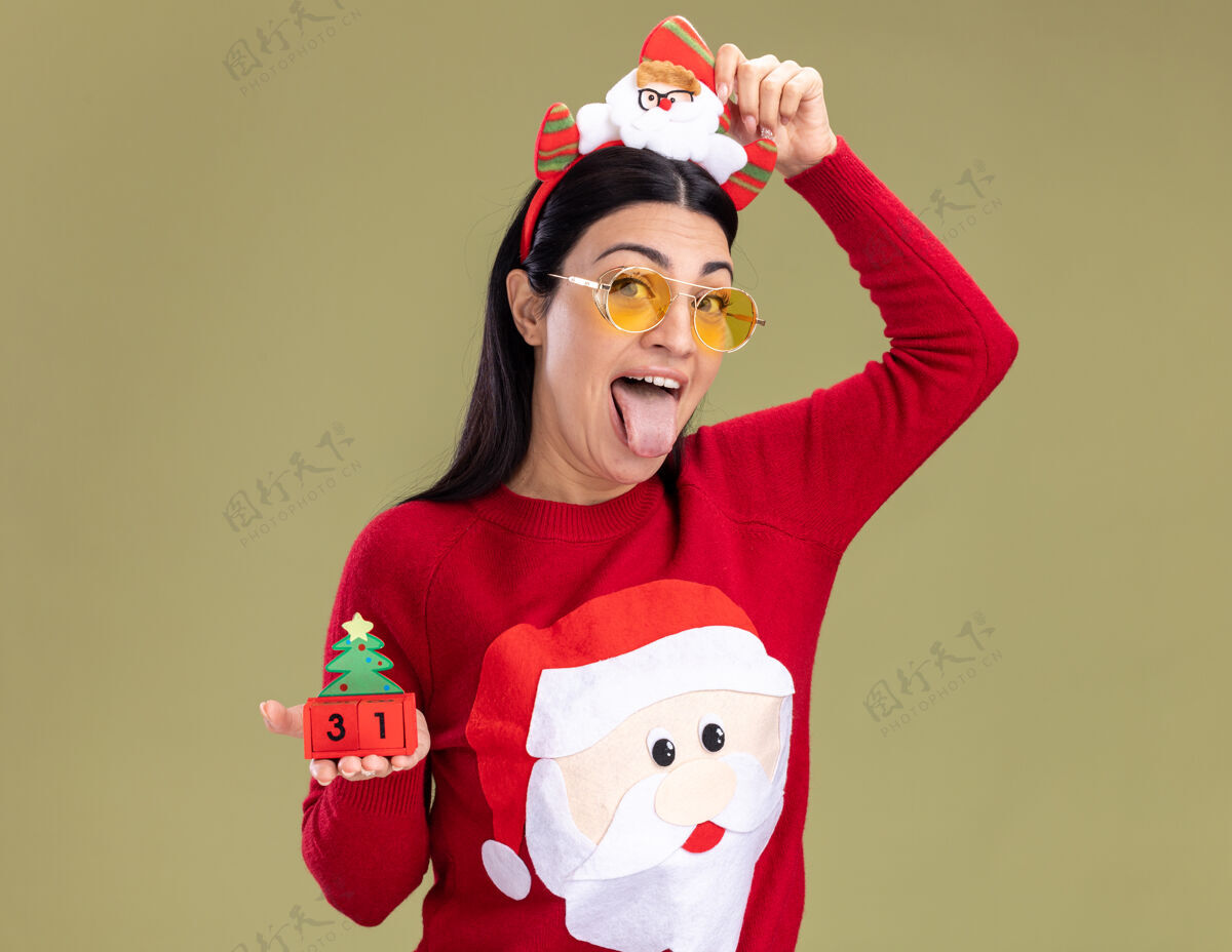 穿顽皮的白人年轻女子戴着圣诞老人的头带和毛衣 戴着眼镜 手里拿着圣诞树玩具头带圣诞快乐圣诞