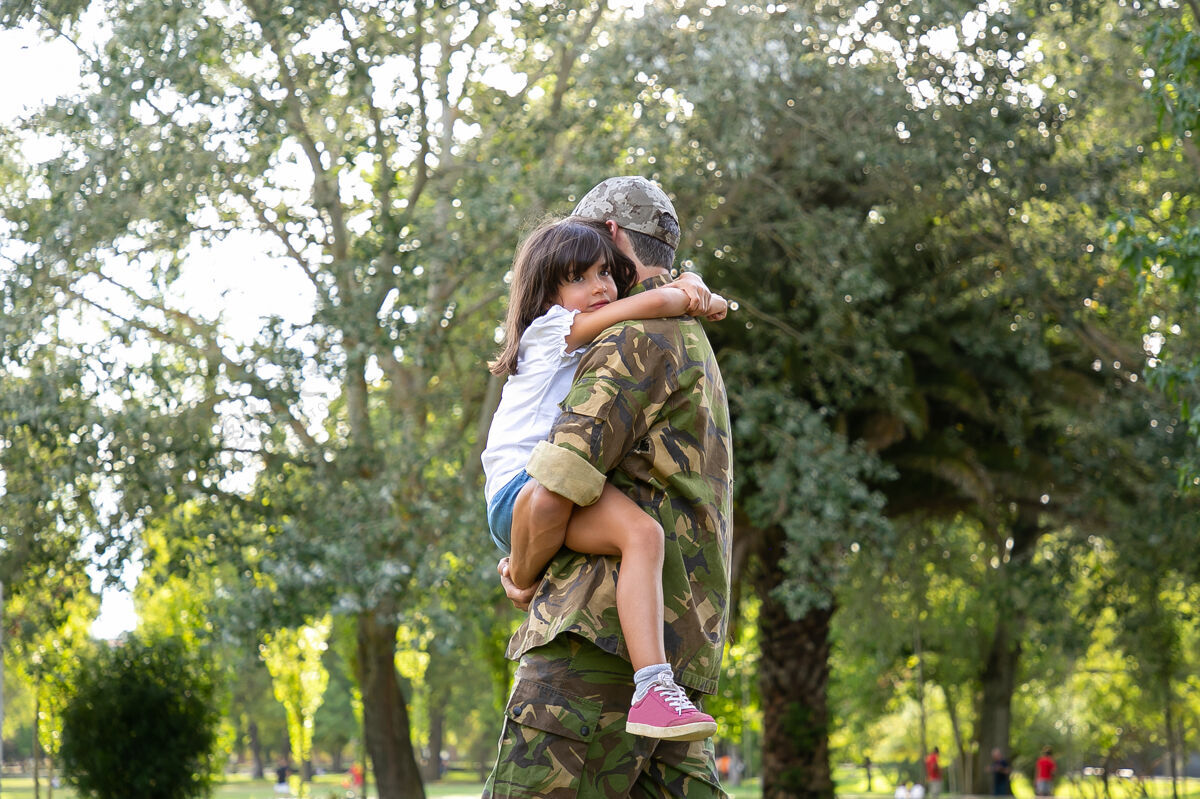 中年穿着军装的白人爸爸抱着女儿中年爸爸站在城市公园里可爱的女孩坐在他的手上 搂着爸爸的脖子童年 周末和军人父母的概念朋友回归脖子