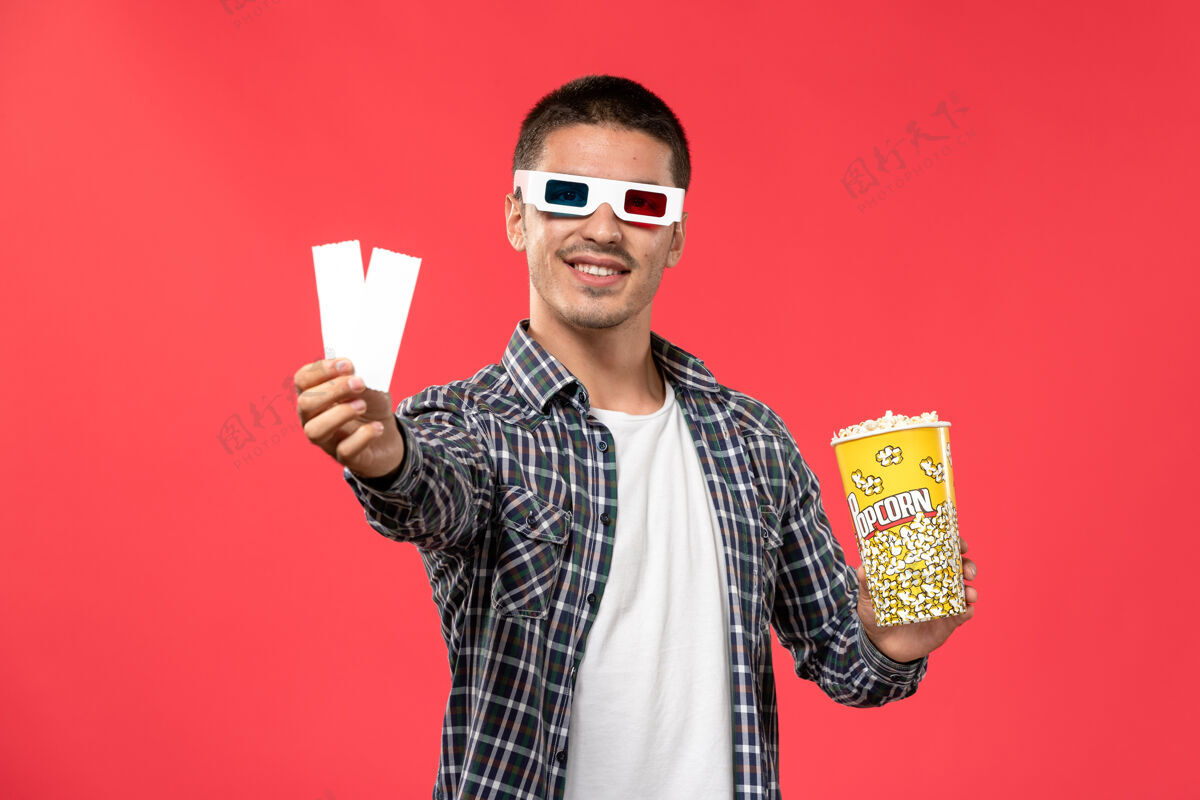 男性正面图年轻男子戴着d型太阳镜 手里拿着爆米花包和电影票 站在浅红色的墙上看电影包装年轻男性电影院