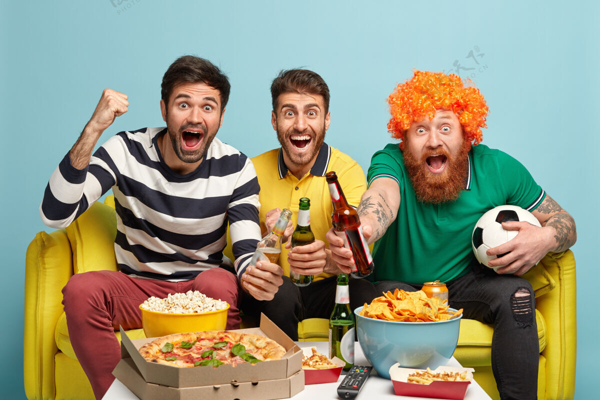 比赛快乐的三个人在周末一起看足球比赛 庆祝进球 坐在黄色沙发上 隔着蓝色的墙人们 兴奋的概念尖叫高兴胡须