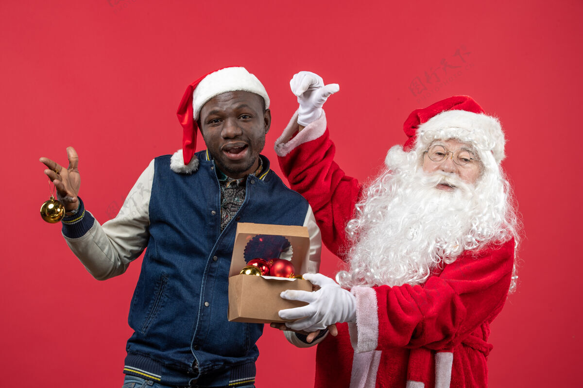 玩具红墙上的圣诞老人和拿圣诞树玩具的年轻人的正视图圣诞老人年轻节日