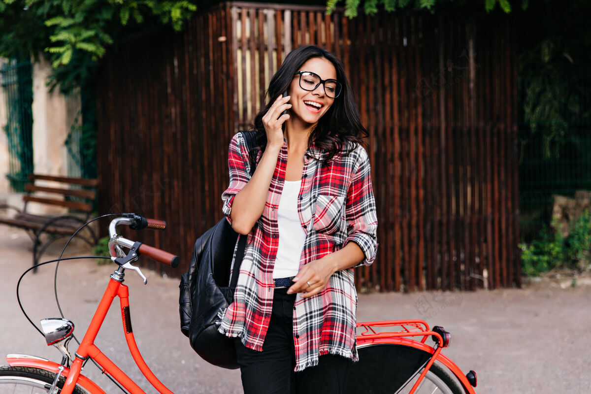 卷发快乐的女人背着黑色的皮包在街上打电话优雅的黑发女孩站在红色的自行车旁边户外夏天微笑