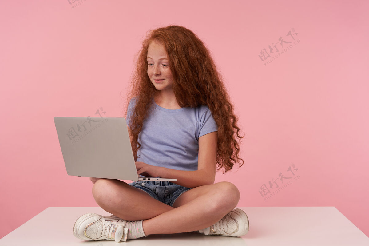 情绪可爱的卷发红发女孩儿 穿着休闲服 盘腿坐在粉色工作室背景上 带着现代笔记本电脑 正眼看着屏幕交叉休闲童年