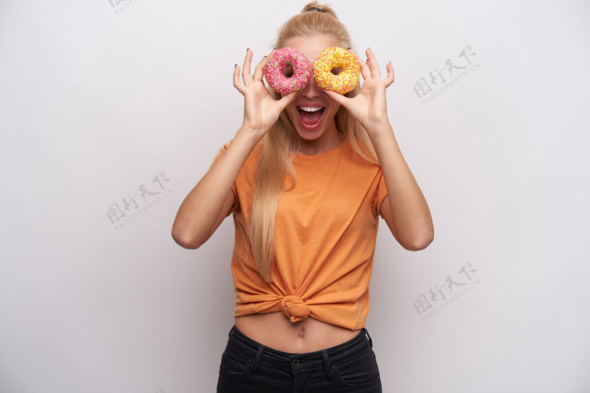 饮食快乐的年轻长发金发苗条女子 站在白色背景下 透过彩色捐款看镜头 开怀大笑抬高穿着食物