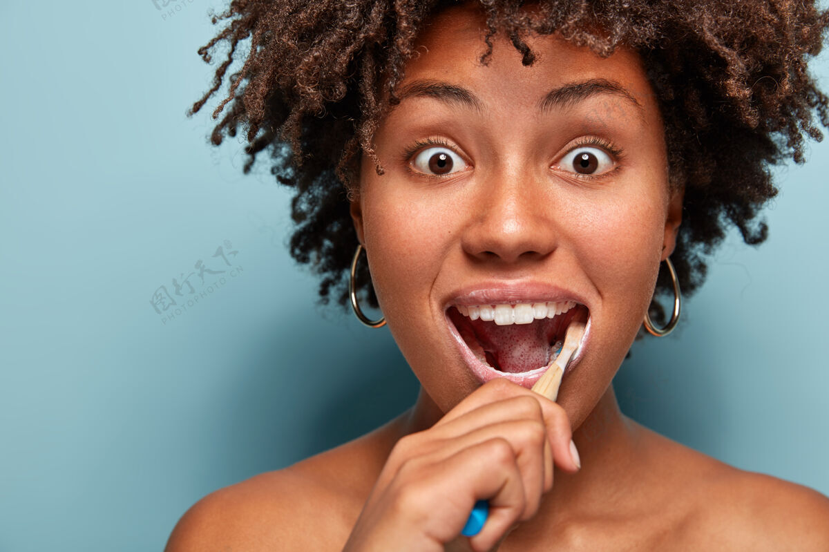 惊讶牙齿卫生和牙科概念头像是一位年轻的非洲裔美国妇女 头发蓬松 用牙刷和牙膏刷牙 眼睛被窃听 眼睛被蓝色隔离卷发洗印象深刻